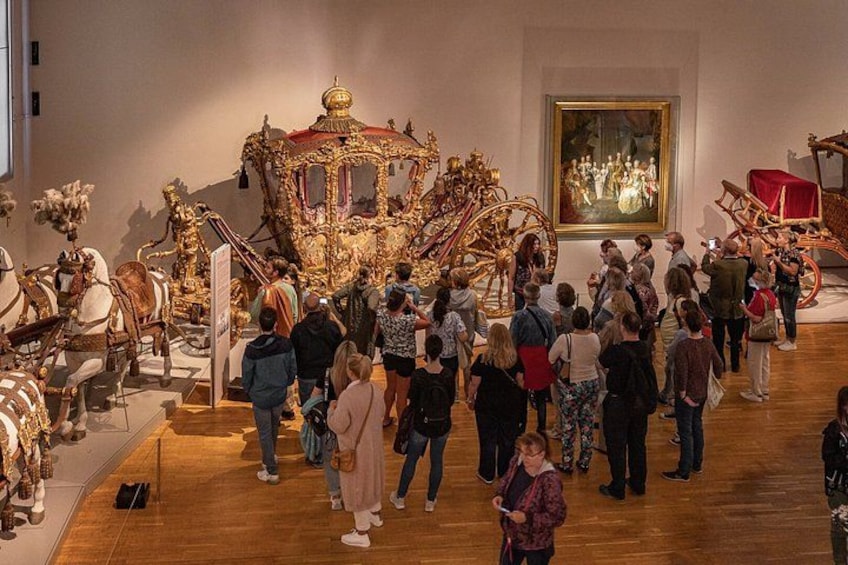 Een kunsttentoonstelling met gedetailleerde gouden koetsen en een groot schilderij aan de muur. 2