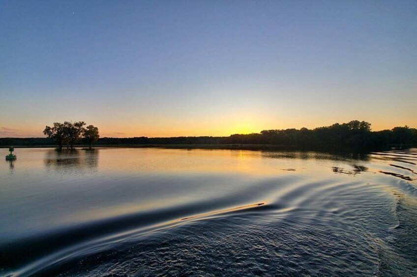 A calm lake at sunset with a boat creating ripples, a tree in the foreground, and a tree line on the horizon. 2