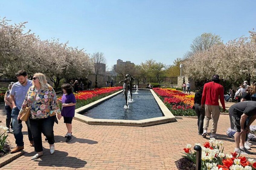 Un parque público con una fuente central, parterres de flores y gente caminando. 2