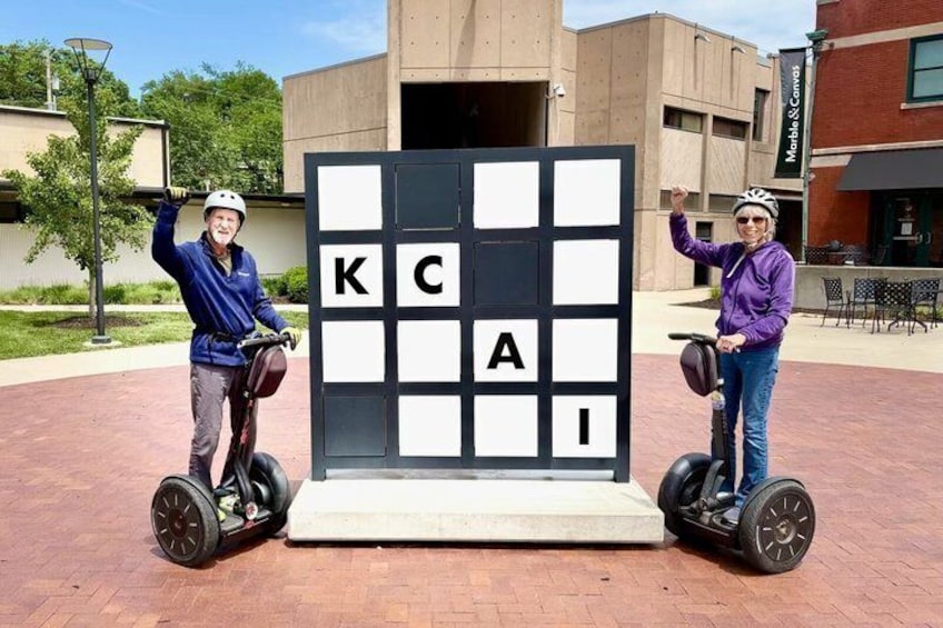 Dos personas en segways frente a un cartel grande con las letras 'KCAI'. 3