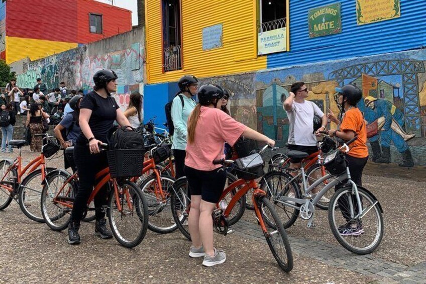 A group of people on bicycles, some wearing helmets, gathered in front of a colorful mural. 5