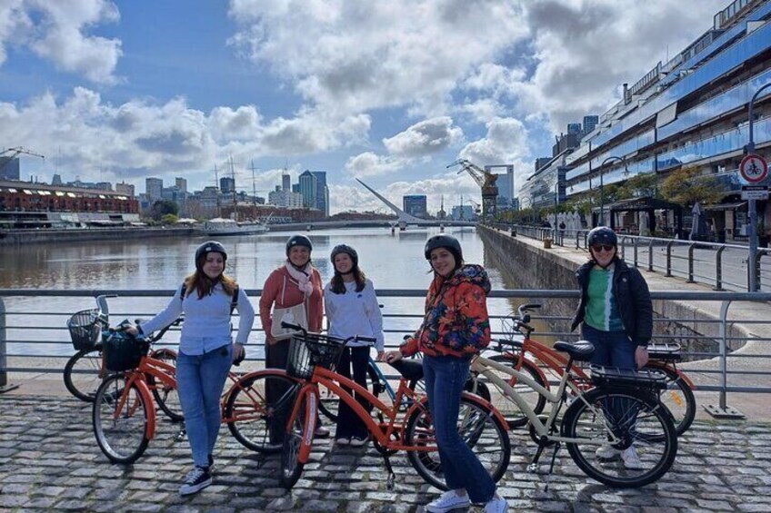 A group of people standing by bicycles on a waterfront promenade with a city skyline in the background. 1