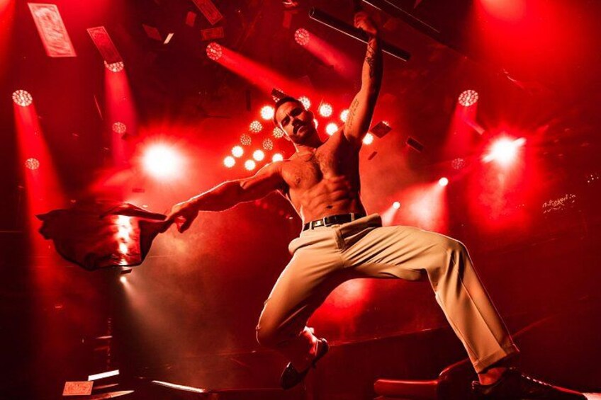 Een man zonder shirt danst in beige broek en houdt een microfoon vast, in een donkere locatie met rode verlichting. 4