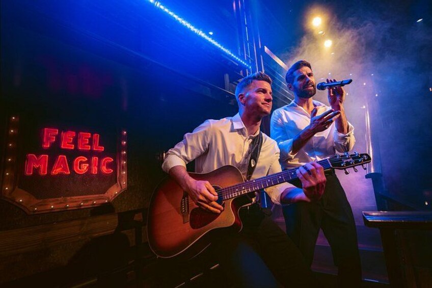 Två personer uppträder med musik på scenen, och ovanför dem hänger en neonskylt med texten ”KÄNN MAGIN”. 4