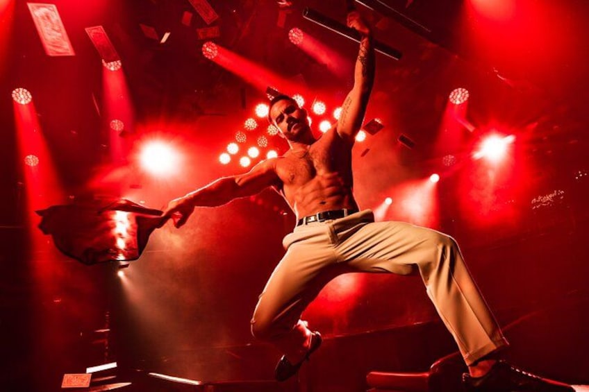 A shirtless man in beige pants performing a dance move with a microphone in a dimly lit venue with red lighting. 4