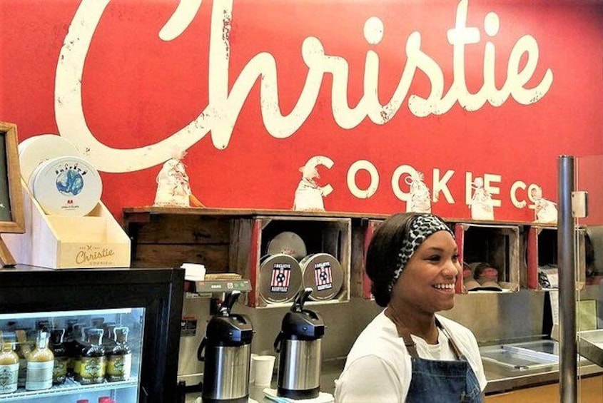 Una donna con camicia e grembiule bianchi in piedi davanti a un muro rosso con le scritte "Christie" e "COOKIES". 3