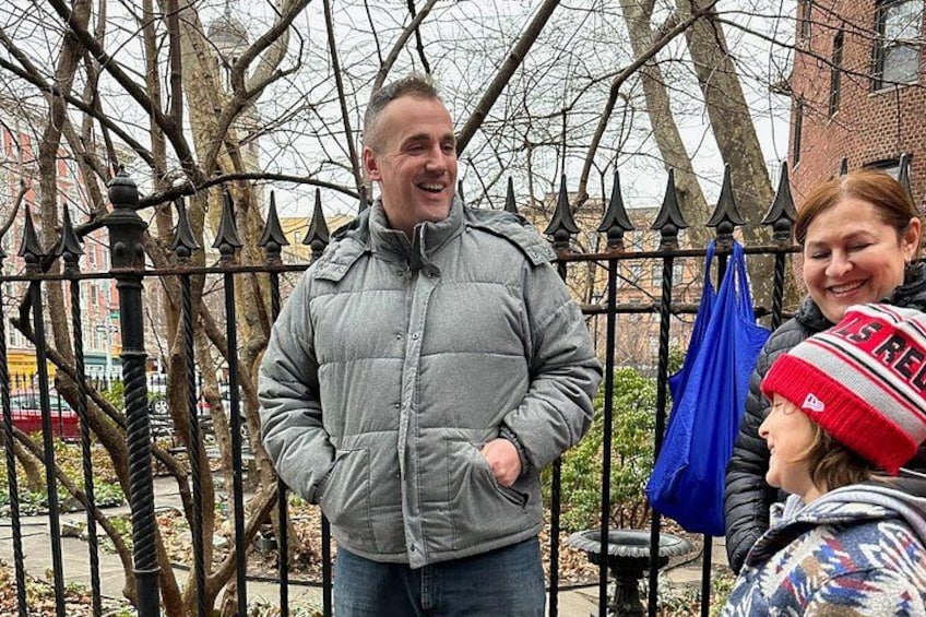 A person in a grey jacket standing in front of a black iron fence with trees and buildings in the background. 5