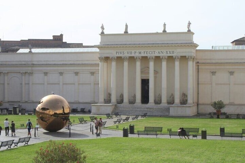 A large golden sphere in a courtyard with classical architecture in the background. 3