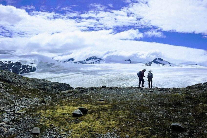 Two people standing on a rocky terrain with a glacier and mountains in the background. 4