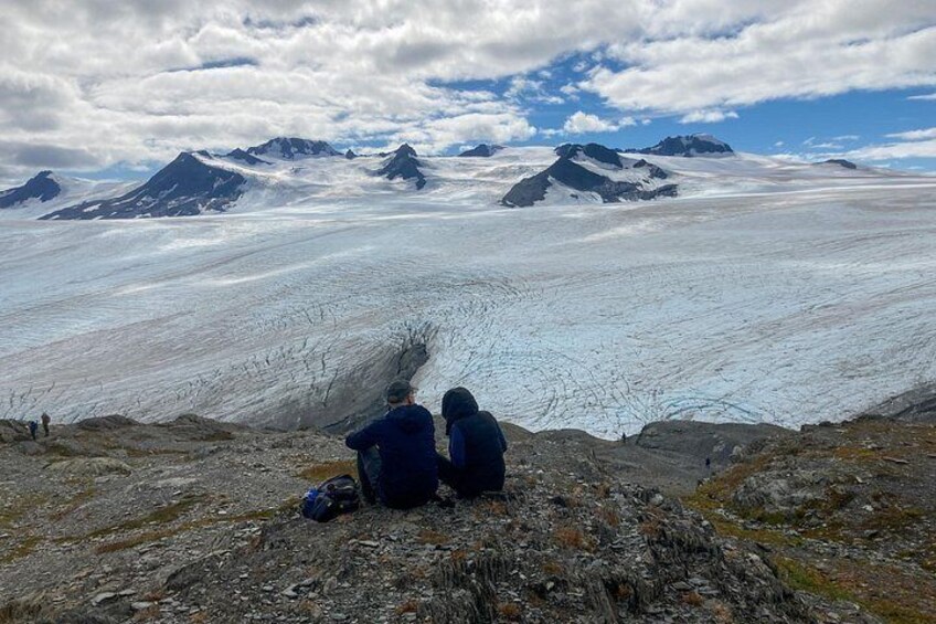 A glacier with two people sitting on a rocky outcrop, mountains in the background, and a cloudy sky. 5