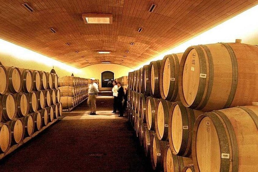 A wine cellar with rows of wooden barrels and a few people inspecting them. 3