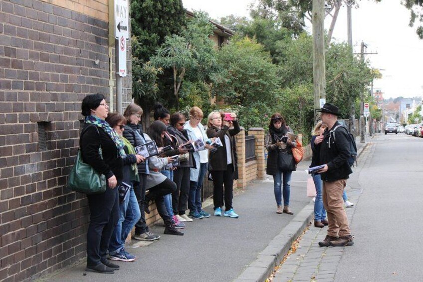 A group of people standing on a sidewalk, some holding papers, with a brick wall and trees in the background. 1