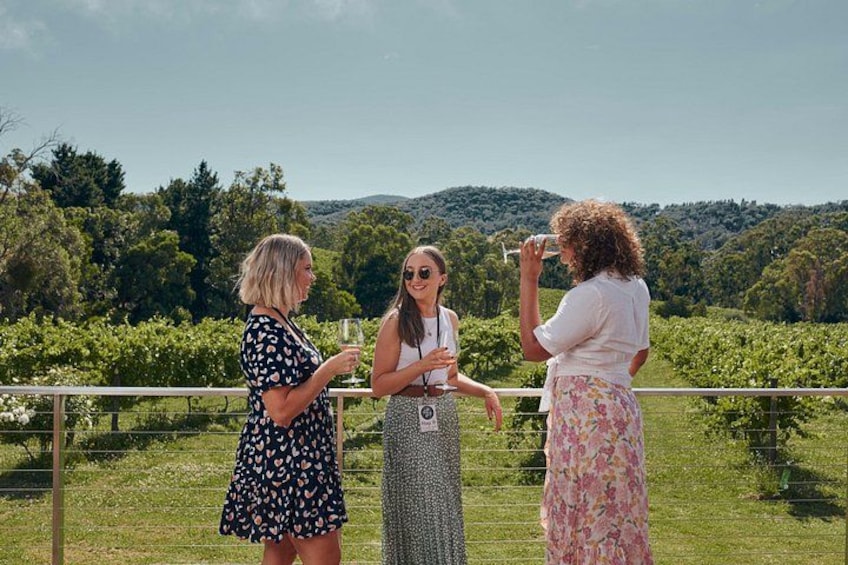 Three people standing outdoors, one holding a camera, another holding a glass, and the third holding a drink, with a vineyard and hills in the background. 1