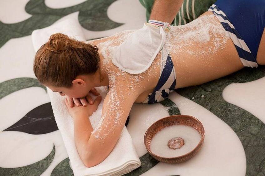En person ligger på ett massagebord, täckt med en handduk, och bredvid finns en skål med vad som ser ut att vara en badprodukt. 2