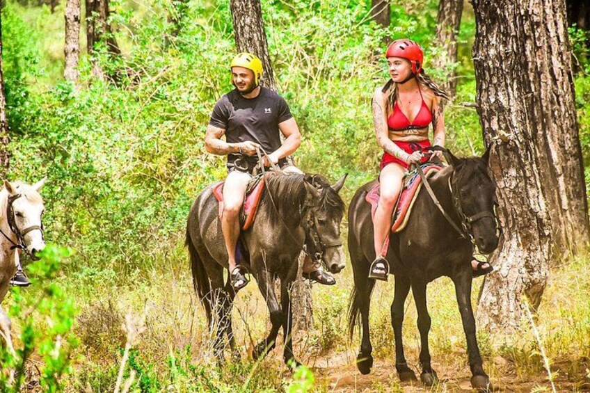 Two people riding horses in a forested area. 5