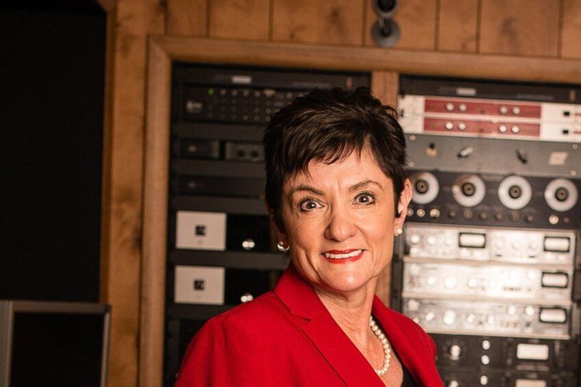 A person in a red blazer standing in front of a control panel with various dials and switches. 3