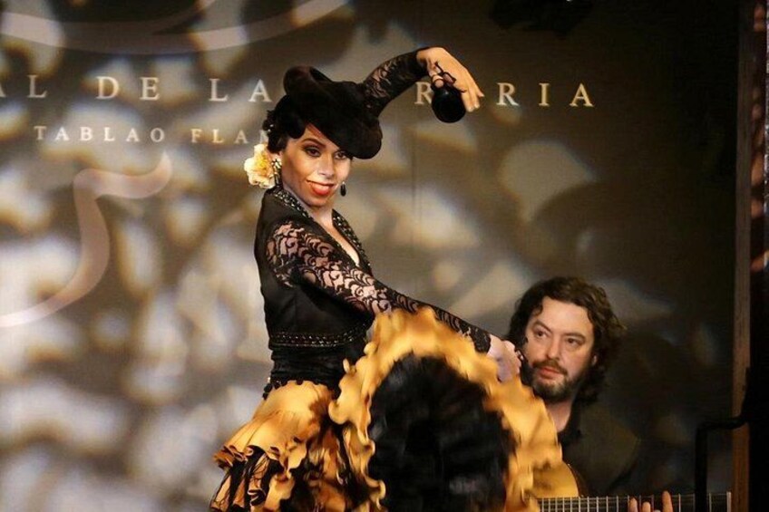 A person in a black and gold outfit with a hat, standing on a stage with a backdrop that reads 'AL DE LA FERRARIA.' 2