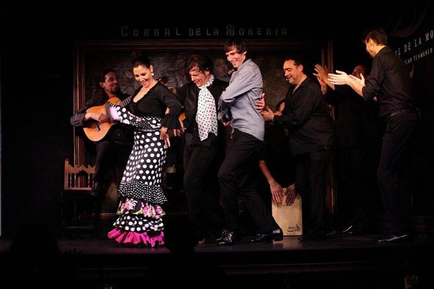 Een groep mensen op een podium met een gitaar, een vrouw in een stippenjurk, en een bord met de tekst 'CORRAL DEL MORERIA'. 2