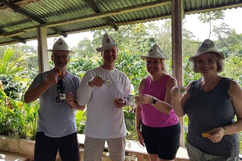 Four people standing outdoors under a shelter, holding drinks, with greenery in the background. 3