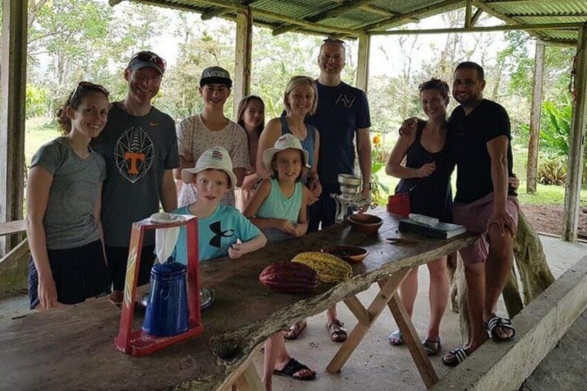 A group of people gathered around a wooden table with a red and blue object on it, possibly a water filter, in an outdoor setting with a covered structure. 2