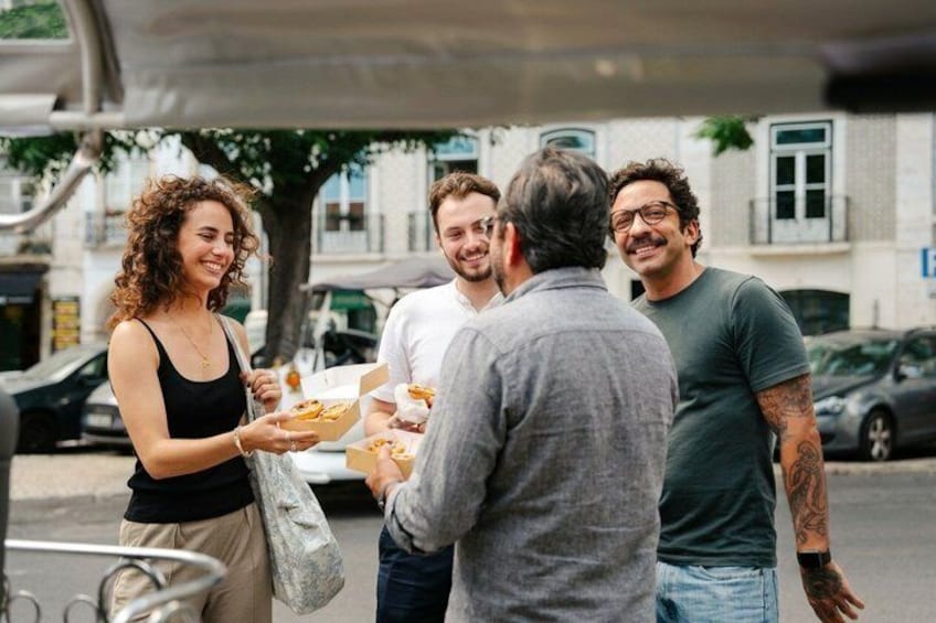 Eine Gruppe von Menschen draußen, einige halten Essensbehälter, im Hintergrund Gebäude und parkende Autos. 2