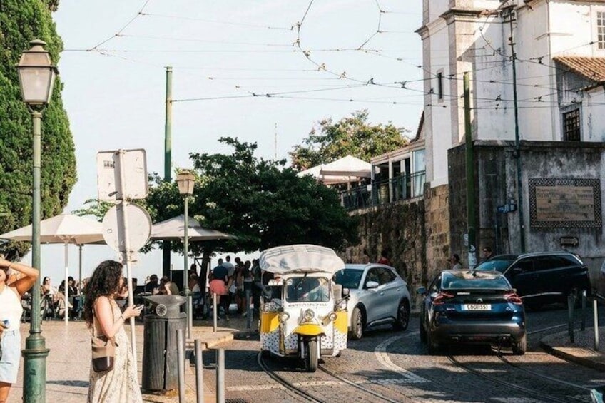 Eine Straßenbahn fährt eine Straße, an der sich Menschen, Autos, Gebäude und Bäume befinden. 3