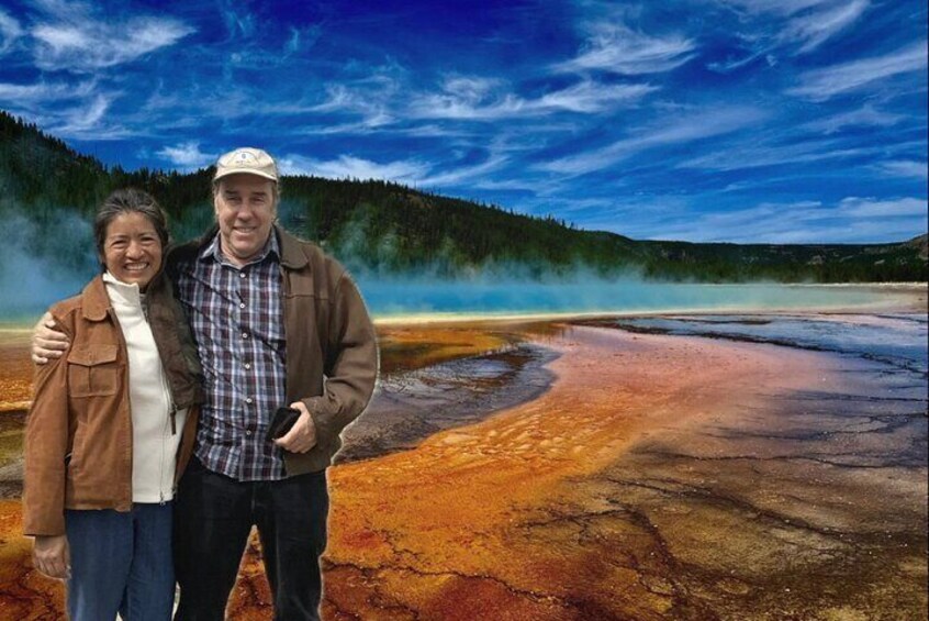 A couple standing in a geothermal area with steam rising from the ground, surrounded by a forested landscape. 1
