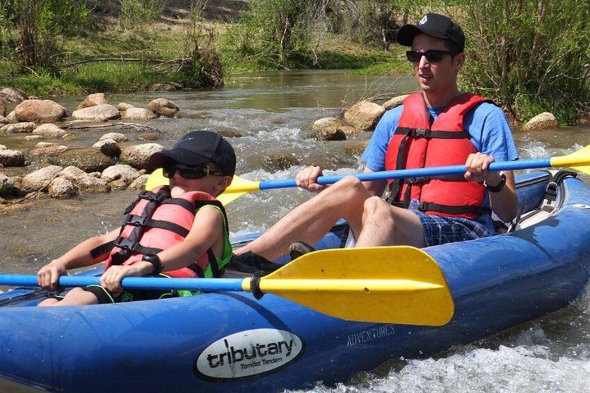 Two people in a blue inflatable raft on a river with rocks. 2