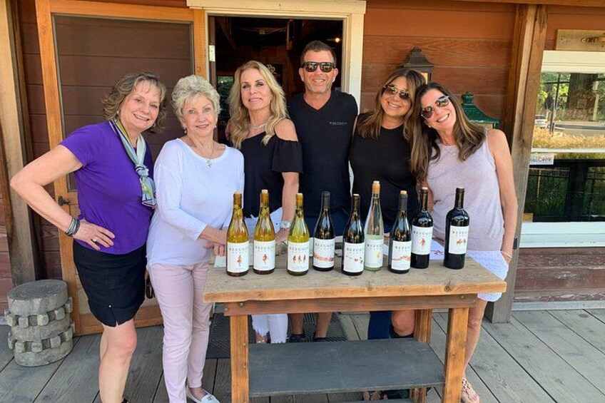 Un grupo de personas de pie frente a una mesa de madera con botellas de vino. 3