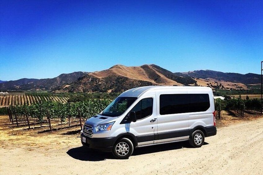 Una camioneta blanca estacionada en un camino de tierra, con viñedos y montañas al fondo. 4