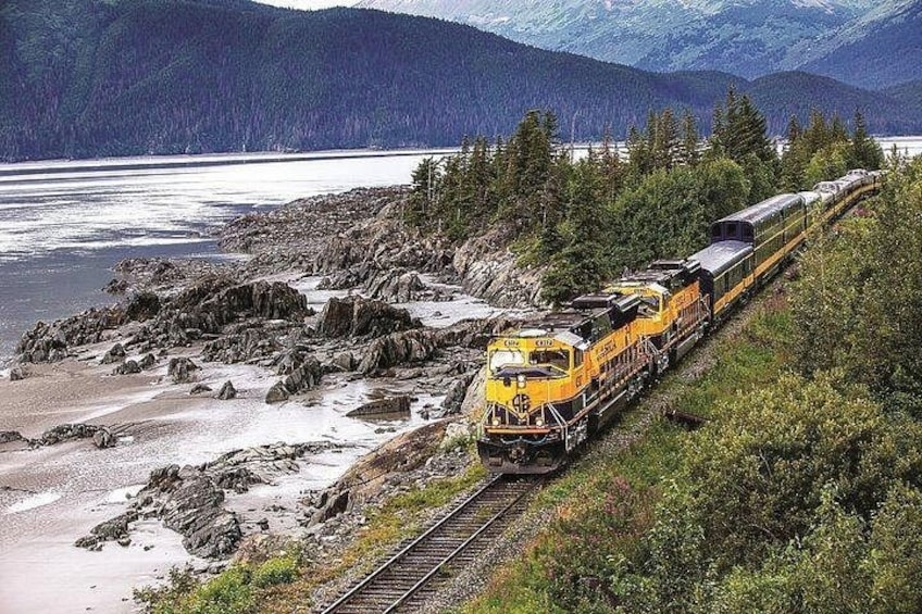 Un tren amarillo sobre rieles cerca de una costa rocosa con un lago y montañas al fondo. 1