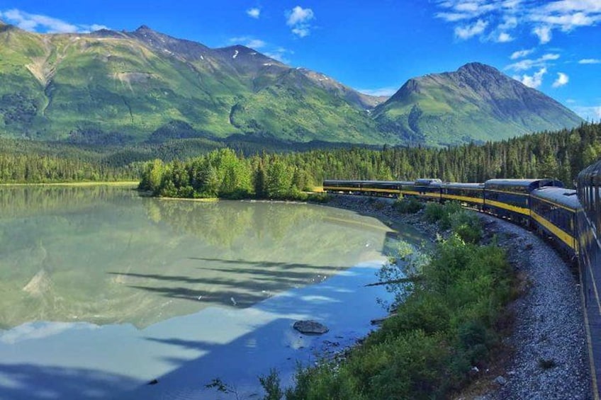 Un tren sobre rieles junto a un lago con montañas al fondo. 2