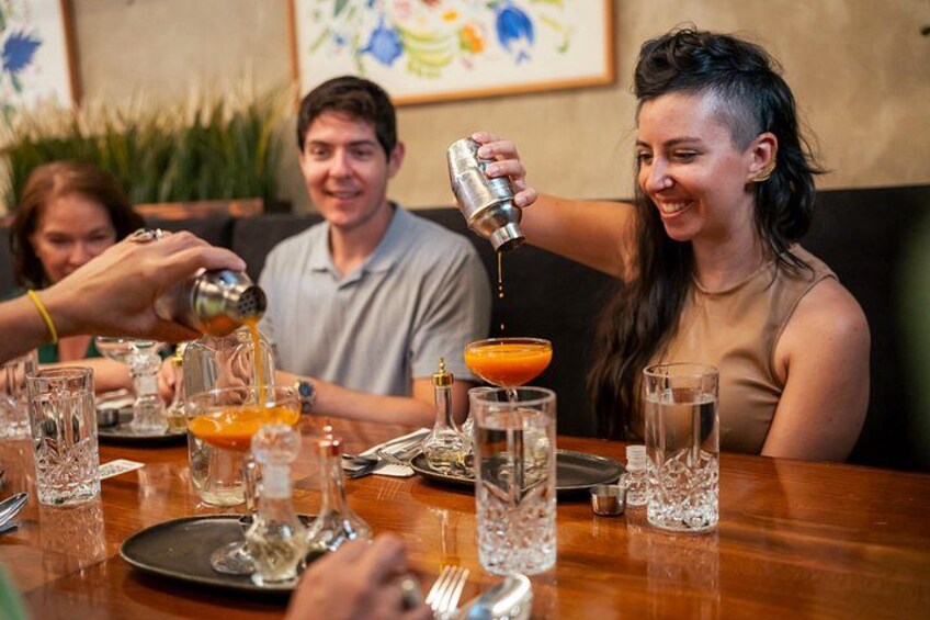 A person pouring a drink into a glass, a table with various glasses and a bottle, and a group of people sitting at the table. 5