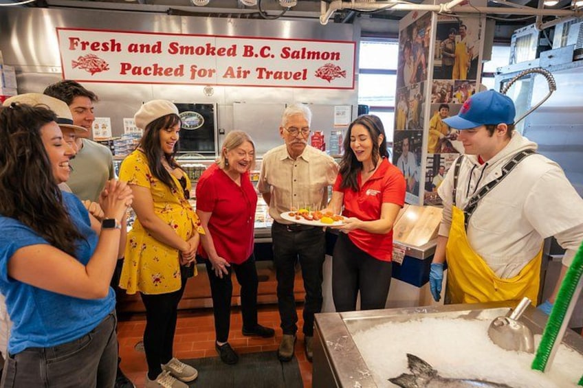 A group of people in a food preparation area with a sign reading 'Fresh and Smoked B.C. Salmon Packed for Air Travel.' 1
