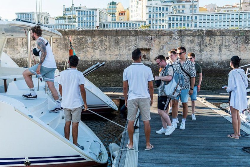 En grupp människor på en brygga med båtar, en stadsbild i bakgrunden och en stenmur. 2