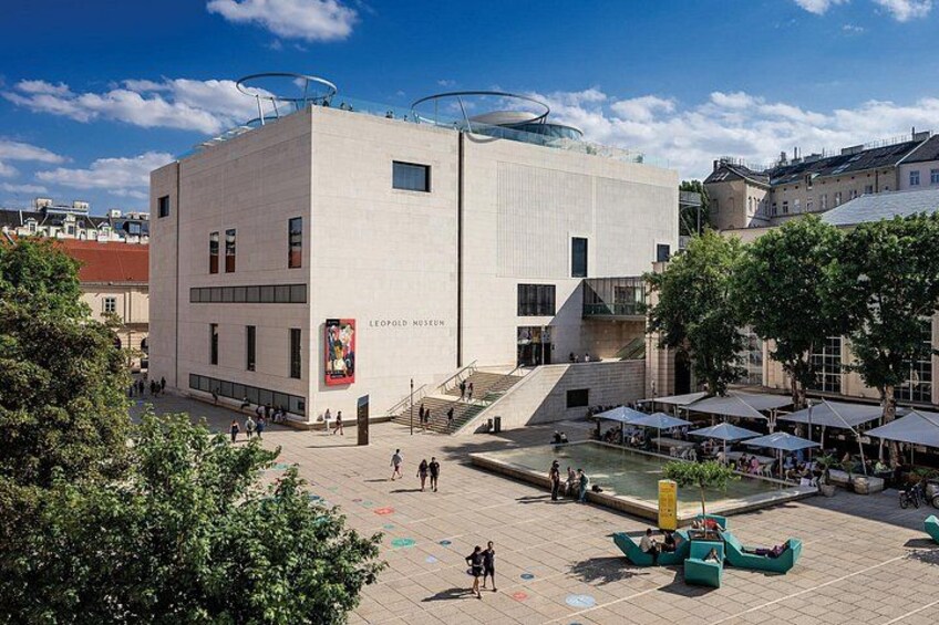 Un edificio moderno con un letrero del MUSEO LEOPOLD, una plaza pública con una fuente y zonas de estar al aire libre. 5