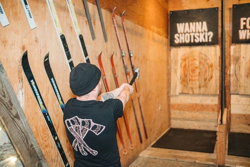 Eine Person zielt mit einem Bogen auf ein Ziel in einer hölzernen Innenraumgestaltung, während Ski an der Wand lehnen und Schilder mit der Aufschrift 'WANNA SHOTSKI?' zu sehen sind. 2