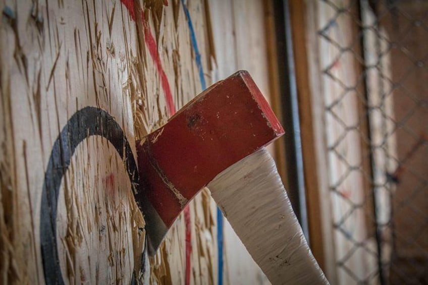 A close-up of a wooden target with a red and white axe embedded in it. 1