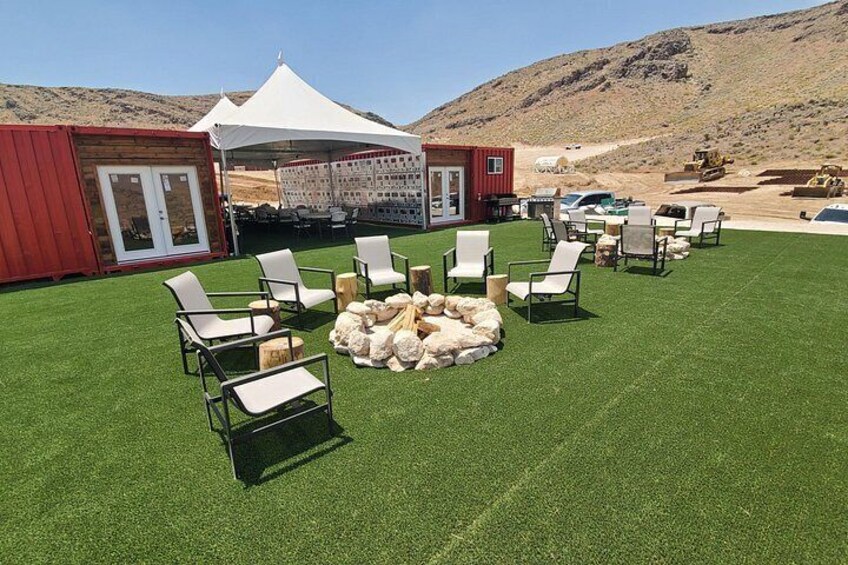 Un área de estar al aire libre con césped artificial, sillas blancas y un hogar para fuego. 3