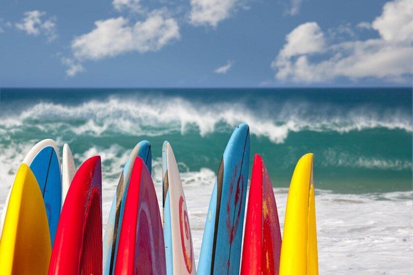 Una hilera de tablas de surf coloridas en una playa, con olas al fondo. 1