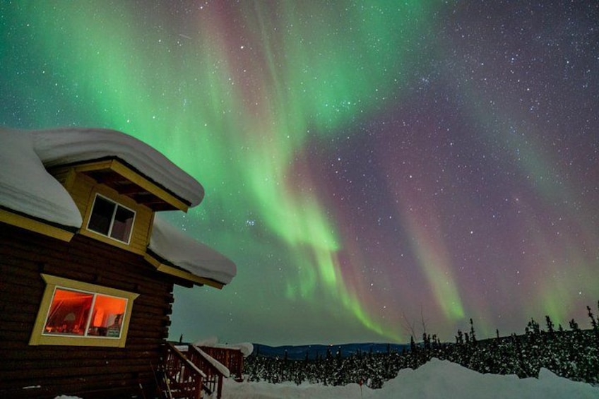 En stuga byggd av trä med ett snötäckt tak, ett snöigt landskap och norrsken på himlen. 1