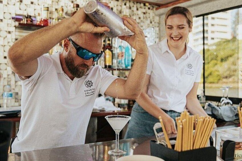 Una persona sirve una bebida en un vaso, otra persona está detrás, y se observa una barra con varias botellas y una copa de martini. 2