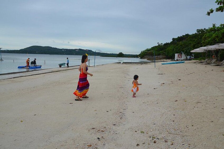 A beach with a woman in a colorful dress and a child walking on the sand, a body of water, and a distant hill. 3