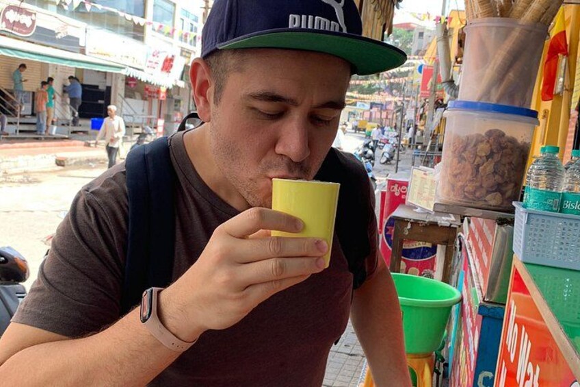 Une personne tenant une tasse jaune, portant une casquette et une montre, se tient dans une rue animée bordée de boutiques, avec un conteneur vert en arrière-plan. 3