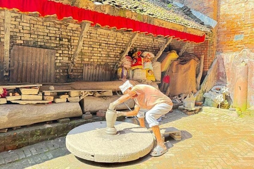 Un uomo che aziona una ruota idraulica tradizionale, un edificio in mattoni con tenda rossa e una statua sullo sfondo. 3