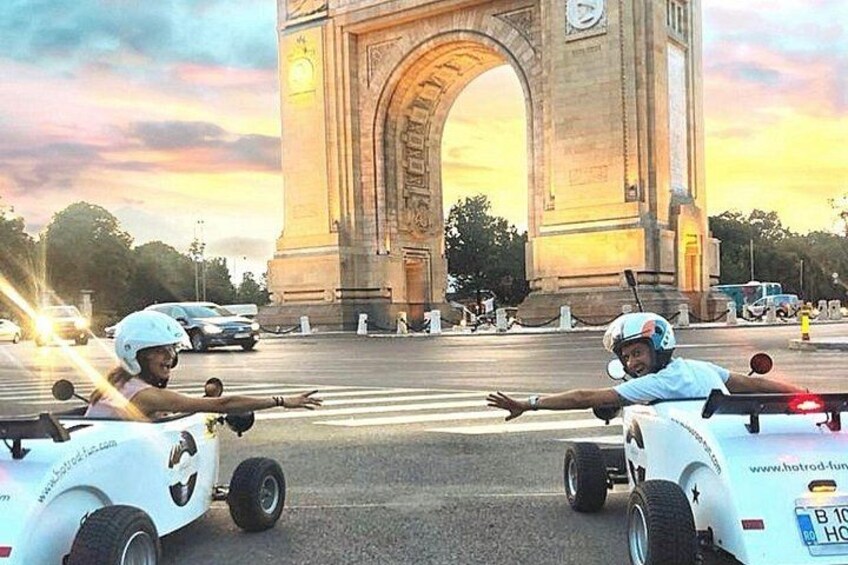 Two individuals in racing cars with helmets, a large arch structure, and a sunset in the background. 4