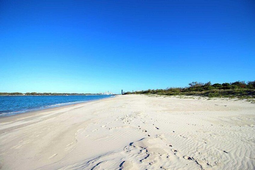 Una playa arenosa con huellas que conducen al agua, un cielo azul despejado y edificios a lo lejos. 2