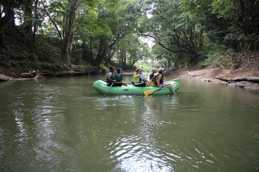 A group of people in a green inflatable raft on a river surrounded by dense trees. 1