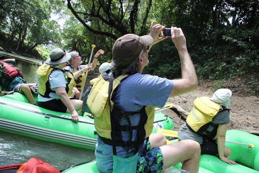 People on a green raft, wearing life jackets and hats, taking a photo. 4