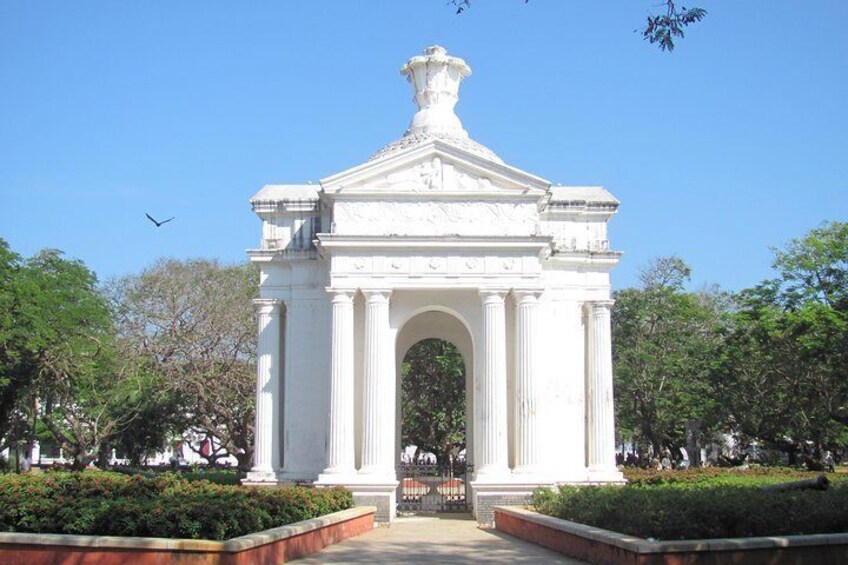 A white neoclassical monument with columns and a central archway, surrounded by well-maintained hedges and trees. 1